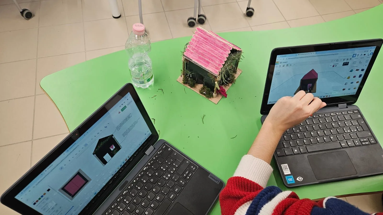 Classroom workshop: on the table, a small house built with natural materials and two laptops showing its 3D model in progress.