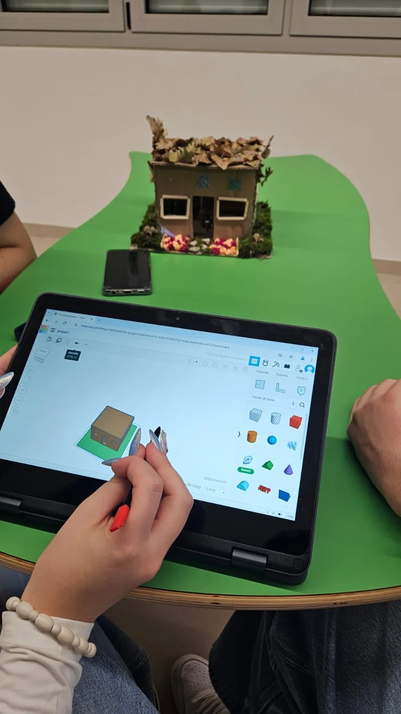 Mani di una studentessa che modella in 3D la casetta costruita con materiali naturali, visibile sul tavolo.